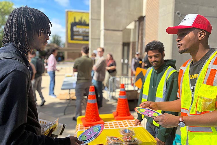 Two black men, one wearing a florescent yellow vest, talks to each other with a table between them. Another young man stands in the background. Two black men, one wearing a florescent yellow vest, talks to each other with a table between them. Another young man stands in the background.