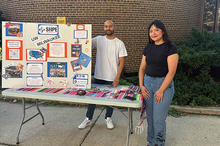 A young man stands behind a table and displays a large poster, while a young women, next to the table, looks are the camera. A young man stands behind a table and displays a large poster, while a young women, next to the table, looks are the camera.