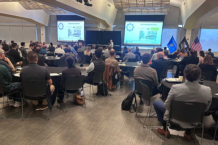 A room full of people listening to a speaker. Two screens in the back display PPT tiles.