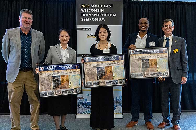 Five people in a row, three men and two women. The middle three are holding award plaques.