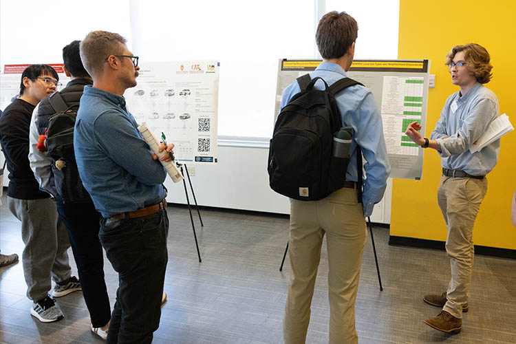 A group of young men stand around a poster listening while the one on the far right talks about it.