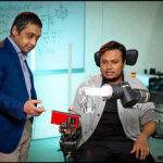 Two men in discussion: One, left is standing & wearing a dark blue jacket and the other is seated in a wheelchair outfitted with a robotic arm.