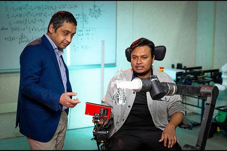 Two men in discussion: One, left is standing & wearing a dark blue jacket and the other is seated in a wheelchair outfitted with a robotic arm.