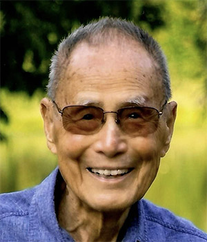 Head shot of an older Asian man, smiling and wearing sunglasses and a light blue shirt.