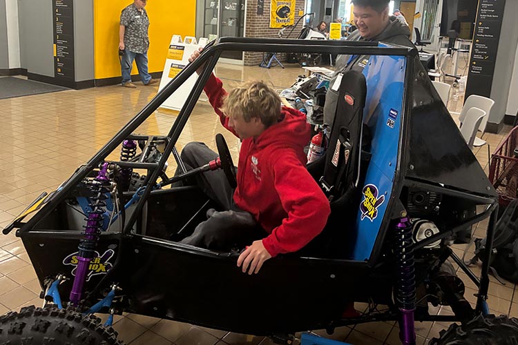 A teen with blonde hair and a red sweatshirt get into the driver's seat of a Baja car.