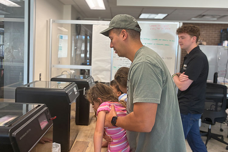 A man in a grey T-shirt raises up a child in pink to see a 3D printer in action.