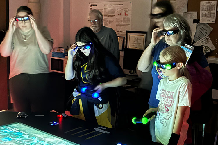 A group of six people, five wearing specialized glasses, learn how to use a holographic table.