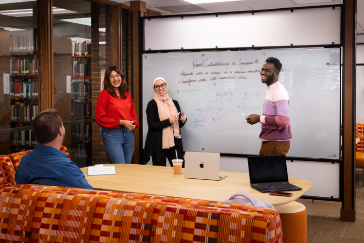 Three international students happily presenting information to a professor. A whiteboard with information from their presentation is behind them.