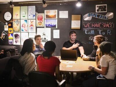 Students sitting around a small, circle table. Some of them are laughing, while others look shocked or stunned.