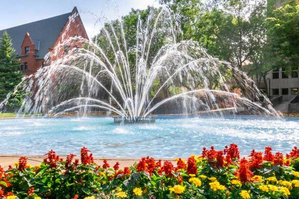View of UWM plaza with a fountain in the middle. It is summer time in the picture, with the trees and flowers in full bloom.