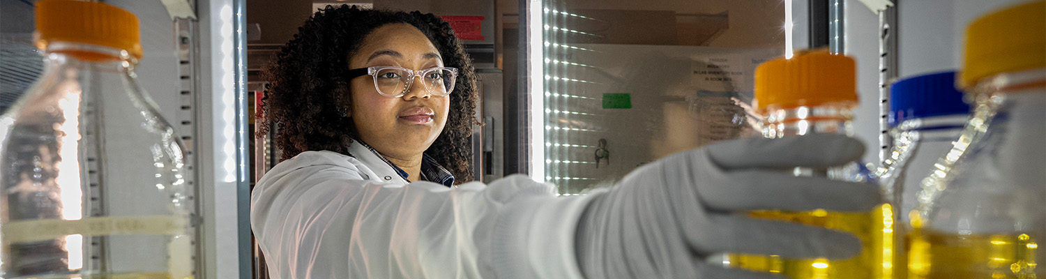 A woman in a lab coat reaches into a fridge for a bottle of yellow fluid.