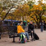 A man and woman sit on a black bench talking to each other. There are changing leaves on the trees behind them and the city bus is driving by in the background.