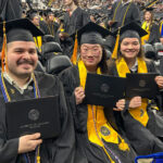 Students display black diplomas while seated at a graduation ceremony.