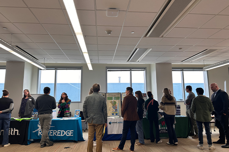 A crowd of people gather by tables talking to each person at the tables.