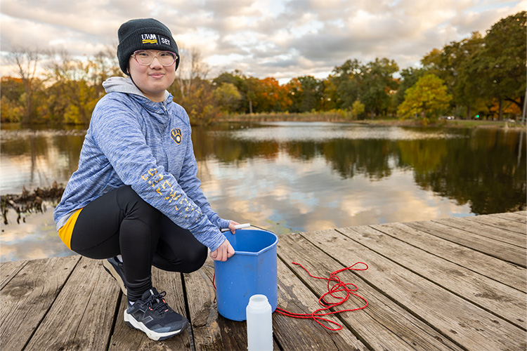 A woman is holding a blue pale of water on a wooden dock with a lagoon behind her. There are trees in the distance.