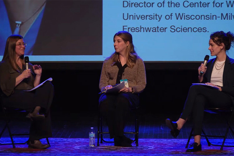 Three women are on a stage talking to each other with a digital presentation behind them.
