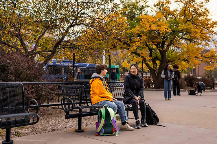 A man and woman sit on a black bench talking to each other. There are changing leaves on the trees behind them and the city bus is driving by in the background.