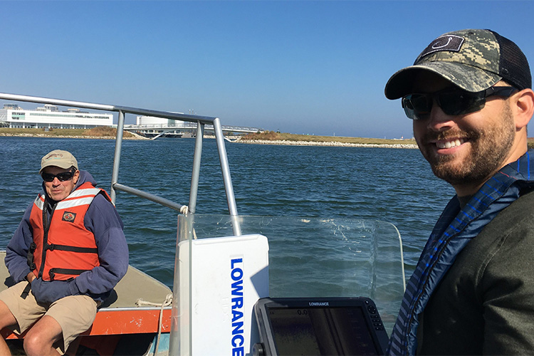 Two men are on a boat smiling. There is blue water in the background.