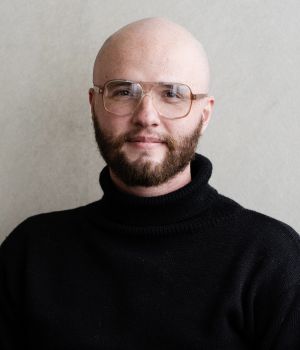Zachary Hying wearing glasses and a black turtleneck against a neutral background.