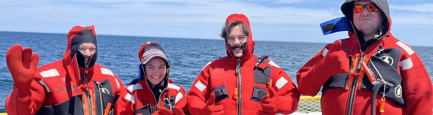 Four people in red immersion suits pose aboard a boat on open water.