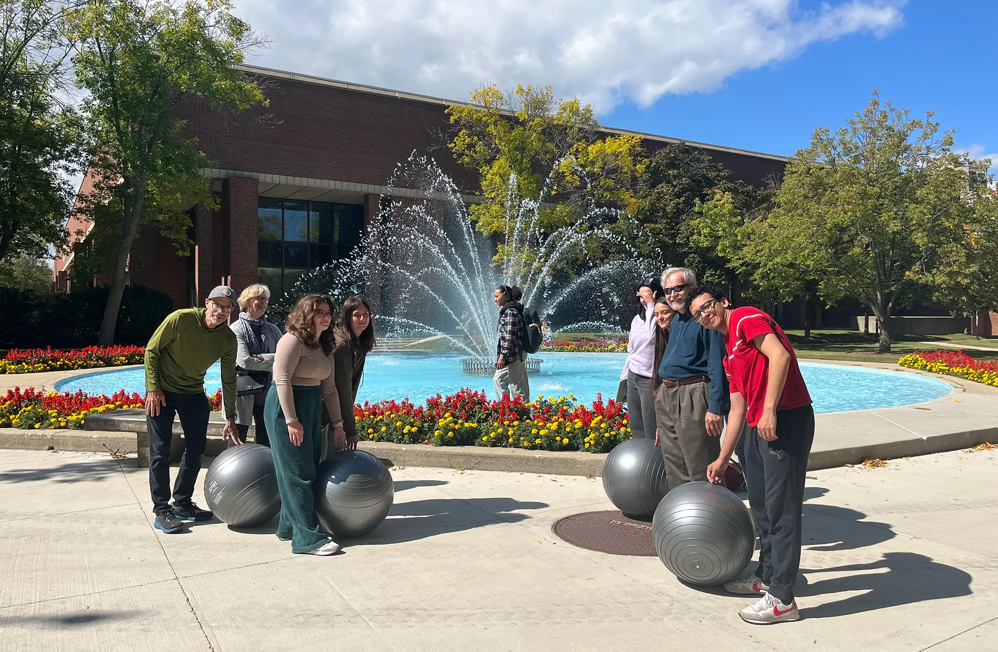 alt text description for photo below: students stand near the fountain beside Curtin Hall outside of Curtin Hall, with large gray yoga balls in lieu of boulders
