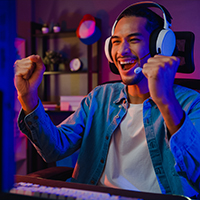 Happy asian guy gamer wearing headphones for participation play video game colorful neon lights computer in living room at night modern house. Esport streaming game online