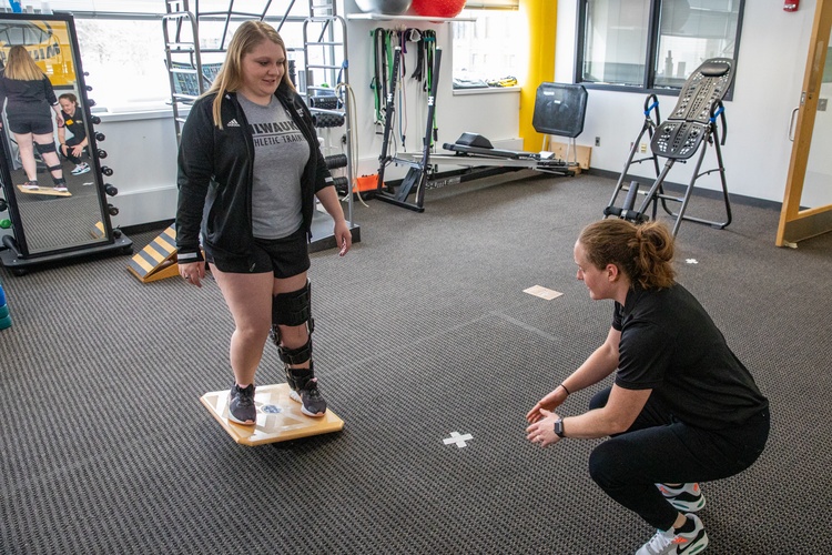MS Athletic Training student assessing the balance and leg strength of a another student using a balance board. The student being assessed is wearing a leg brace.