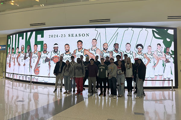 Group of UWM students from the Business Panthers LLC in front of a Fiserv Forum digital display for the Bucks.