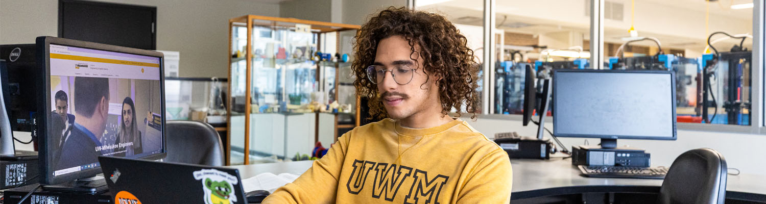 A student wearing a gold UWM sweatshirt works on a laptop in a computer lab. A desktop monitor nearby displays a university webpage, and additional computer stations and equipment are visible in the background. The space appears bright and modern, with large windows and organized work areas.
