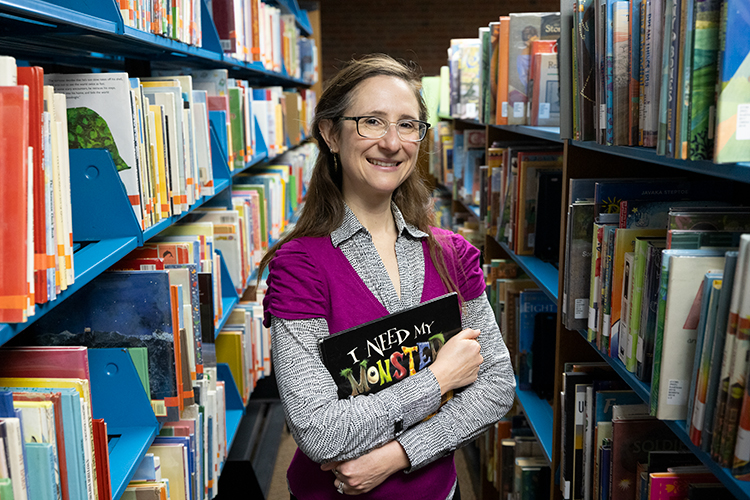 Assistant Professor Sarah Beth Nelson standing in the stacks holding a book between her crossed arms at the UWM library.