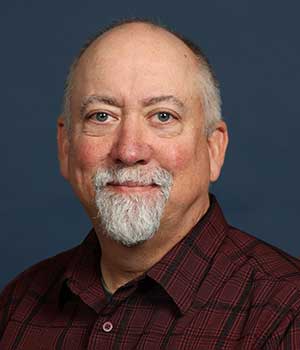 Portrait photo of white male with white beard and mustache. Wearing a red and black dress shirt on a blue background.