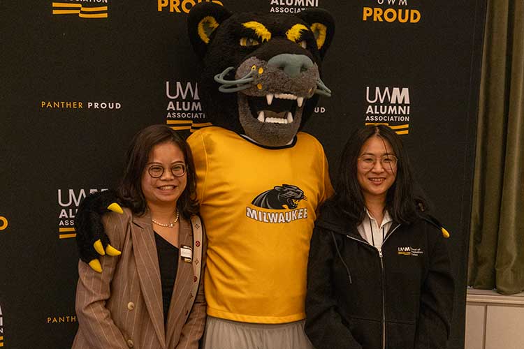PhD Graduate standing in front of a black and gold banner that says UWM Proud. PhD Graduate standing in front of a black and gold banner that says UWM Proud.