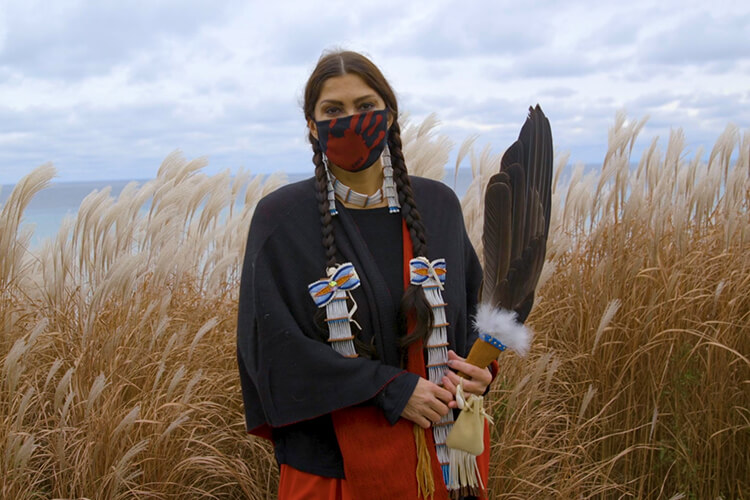 Indigenous woman with facemask with red handprint standing for Missing and Murdered Indigenous Women.  She is standing in front of tall grass at Lake Michigan 