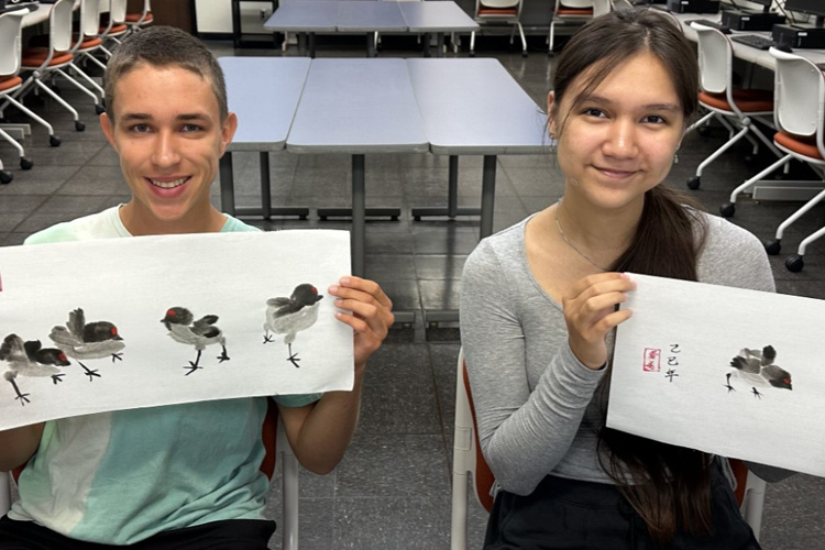 Summer interns sit at a table smiling and holding up their Chinese ink wash paintings of small birds.