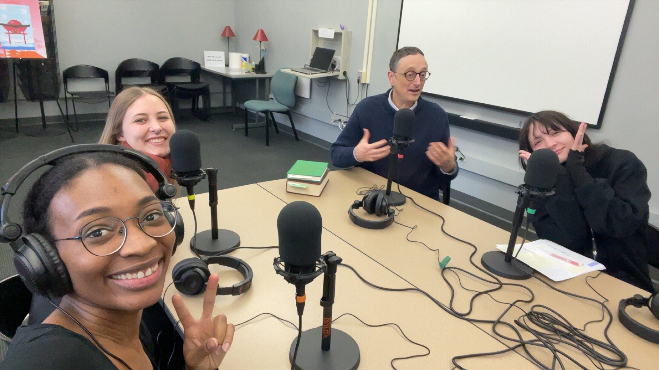 three students and a professor sit at a table in the Language Oasis wearing podcast equipment and smile for a photo