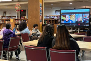 Scene in a school library, with students seated at tables in front of a video screen showing two men on a video call