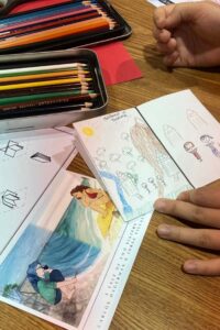 Close up photo of art supplies and two papers: one showing a still frame from the documentary, another showing a student's drawing of landscape and people in Thailand