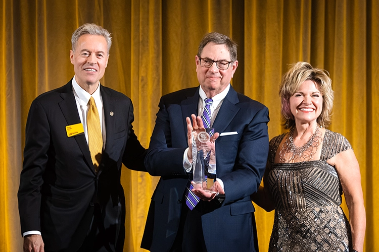 A man in a navy blue suit, striped tie, and glasses, holds a glass award. He stands between UWM Chancellor Mark Mone, wearing a black suit and yellow tie, and UWM Alumni Association Board Chair Michelle Putz, wearing a sparkling evening gown.