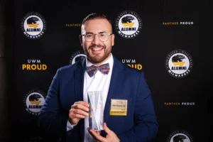 A young man in a navy blue suit and striped bow tie, wearing glasses, holds a glass award in front of a UWM Alumni Association background.