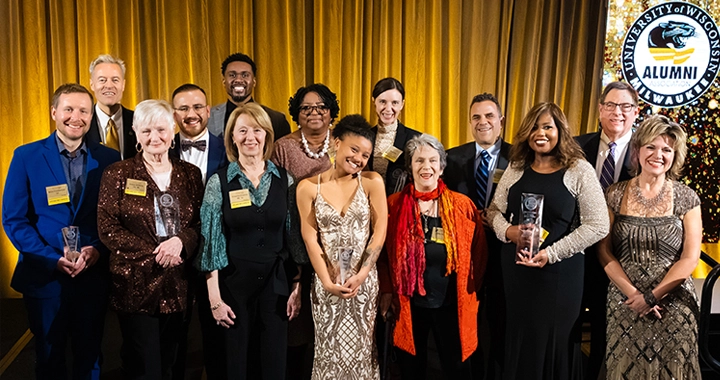 A group of men and women in suits and evening gowns, some holding glass awards, smile in front of a gold curtain background.