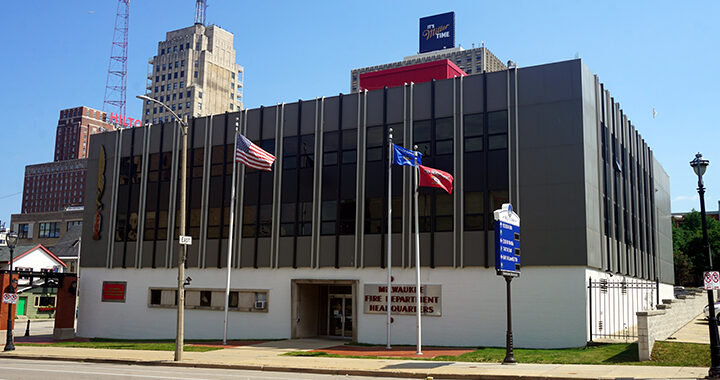 Photo shows the Milwaukee Fire Department headquarters building, a gray facade with vertical windows, a white cinderblock base, and the American and Wisconsin flags in front.