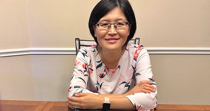 Professor Xiaoxia Cao sits at a table with her arms folded. She is smiling.