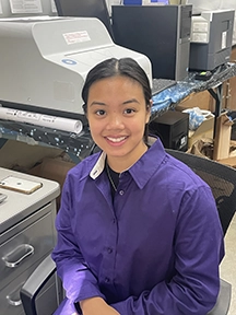 A young woman with dark hair in a purple shirt smiles in front of some laboratory equipment.