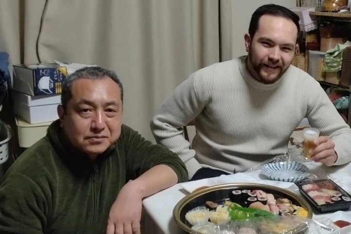 An older Japanese man kneels next to a table, next to a white American man with a beard. Both smile at the camera. The table is set with a spread of sushi.
