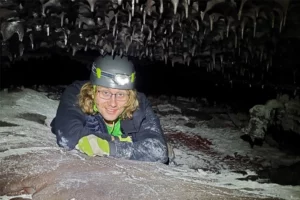 A young white man with shoulder-length blond hair lies on his stomach in an enclosed space. He wears a hard hat and glasses. The low ceiling of the rocky space is covered with small black outcroppings.