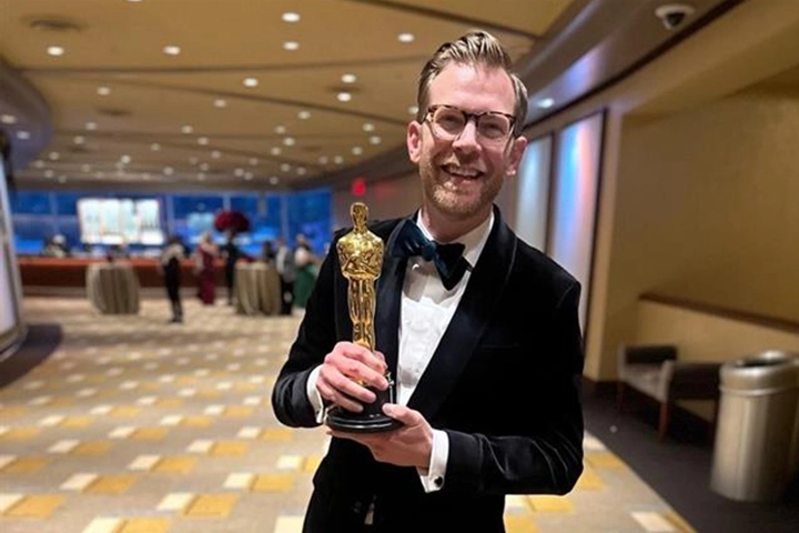 A smiling white man in a tuxedo holds a golden statuette.