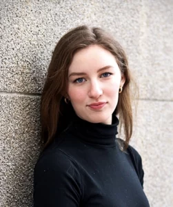 A headshot young white woman with shoulder-length brown hair leaning against the wall of a building.