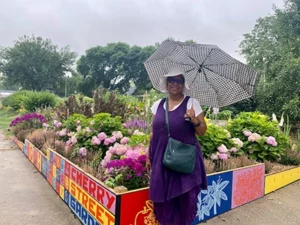 A middle-aged Black woman wears a purple tunic, white hat, and sunglasses. She holds a black-and-white checkered umbrella. She stands in front of a vibrant patch of flowers. A colorful border around the flowerbed reads "Cherry Street Garden."