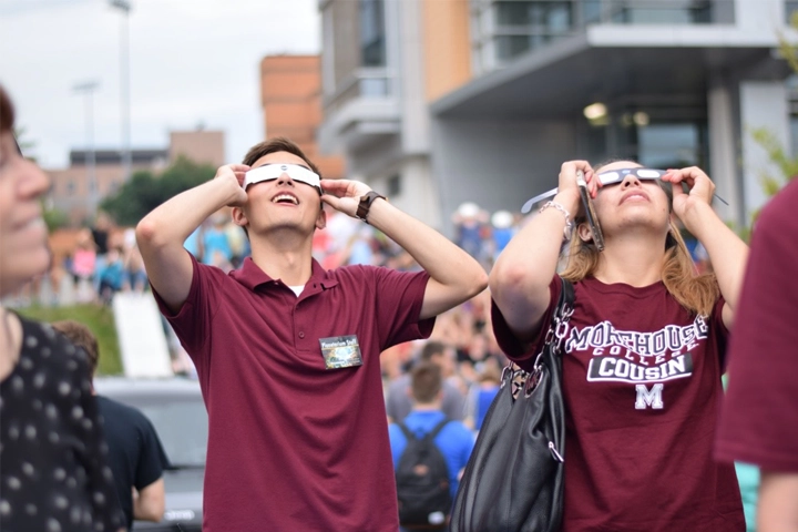 Two young people wearing maroon shirts stare up at the sky while holding paper glasses to their eyes.