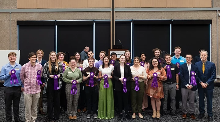 A group of students in business casual clothing stand in a group, all holding purple ribbons.